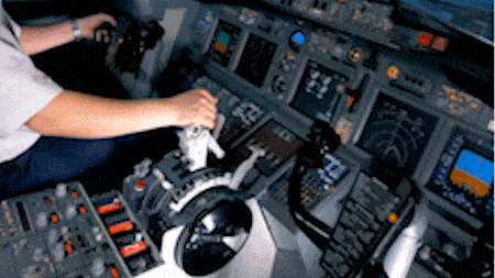 Pilot operating airplane controls in cockpit with instrument panels and screens in background.