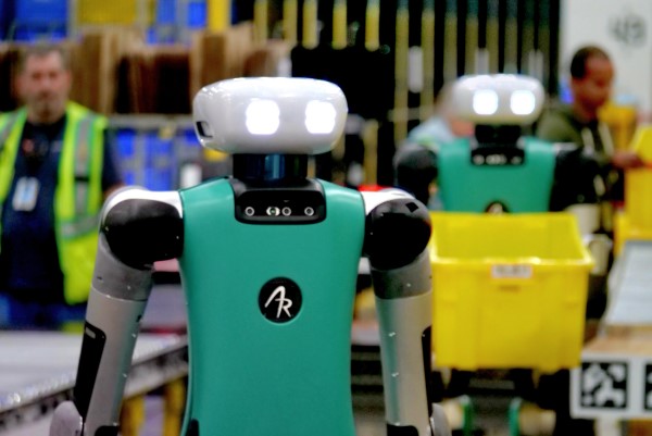 A humanoid robot named Digit, with a teal torso and grey arms, stands in a warehouse. Its white head is lit up, and workers and another robot are visible behind it.