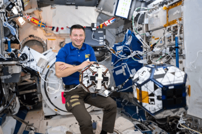 Astronaut in blue shirt holding a geometric robotic device inside a space station module.