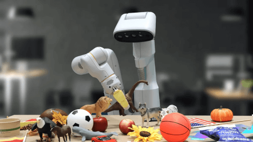 Robotic arm surrounded by learning materials including plastic animals, sports balls, and seasonal decorations on a classroom desk, demonstrating interactive teaching tools.